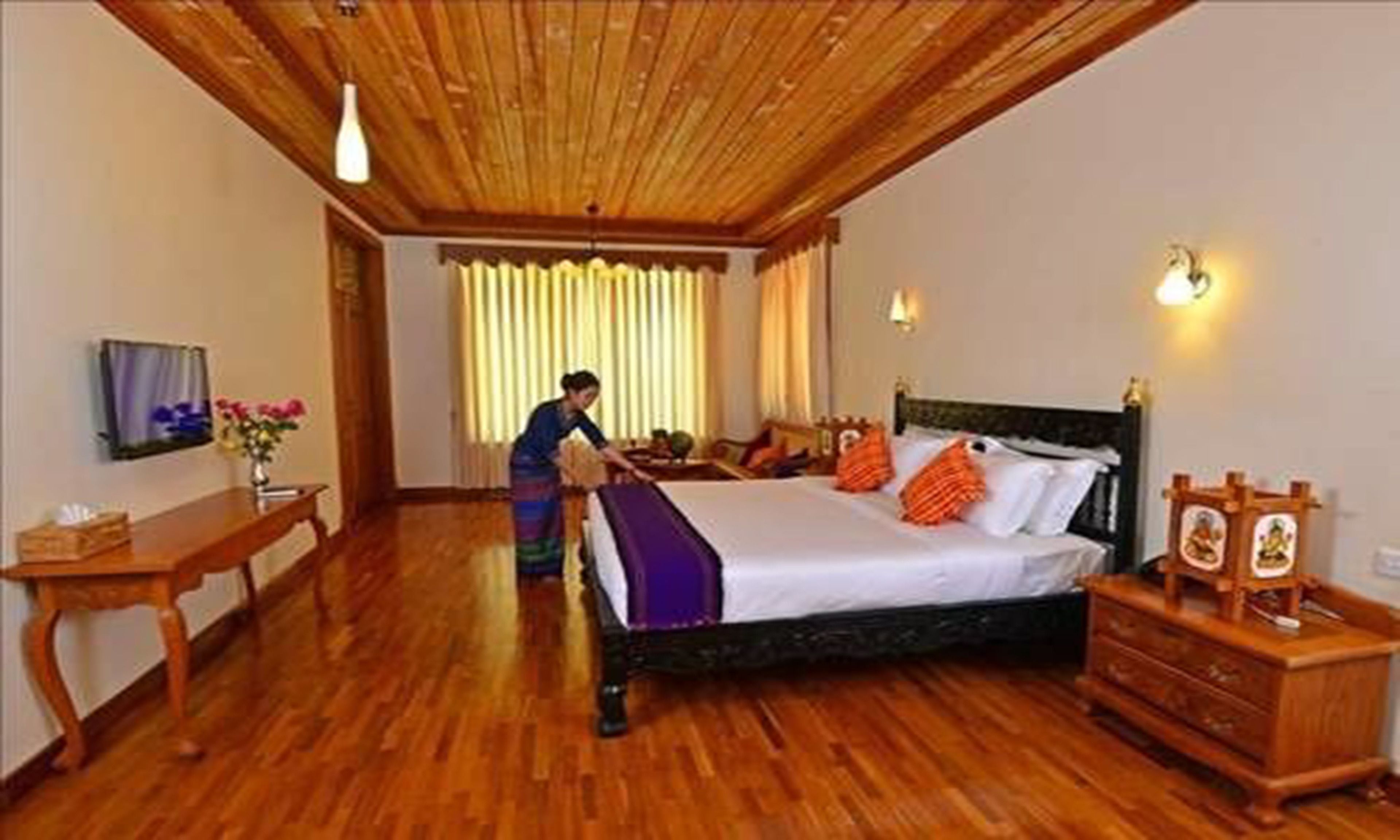 Habitación de hotel con techo de madera, una cama con almohadas naranjas, un aparador de madera y un escritorio con un jarrón de flores.