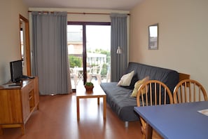 Uma sala de estar com sofá, mesa de madeira, televisão em um móvel e varanda com cadeiras.