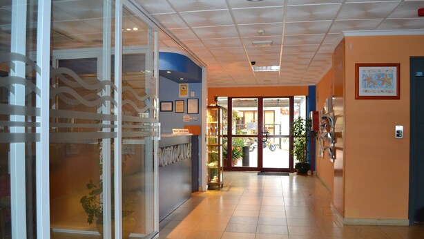 Zona de recepción de un hotel con puerta de cristal de entrada, un mostrador de recepción y un pasillo que lleva a la entrada.
