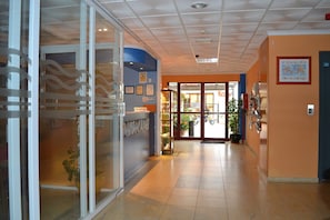 Uma área de recepção de hotel com entrada por porta de vidro, balcão de recepção e corredor que leva à entrada.