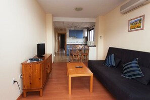 Sala de estar com um suporte de TV de madeira, um sofá preto com almofadas listradas e uma mesa de centro de madeira. Dá para ver uma cozinha ao fundo, com armários azuis, e uma área de jantar com cadeiras de madeira.