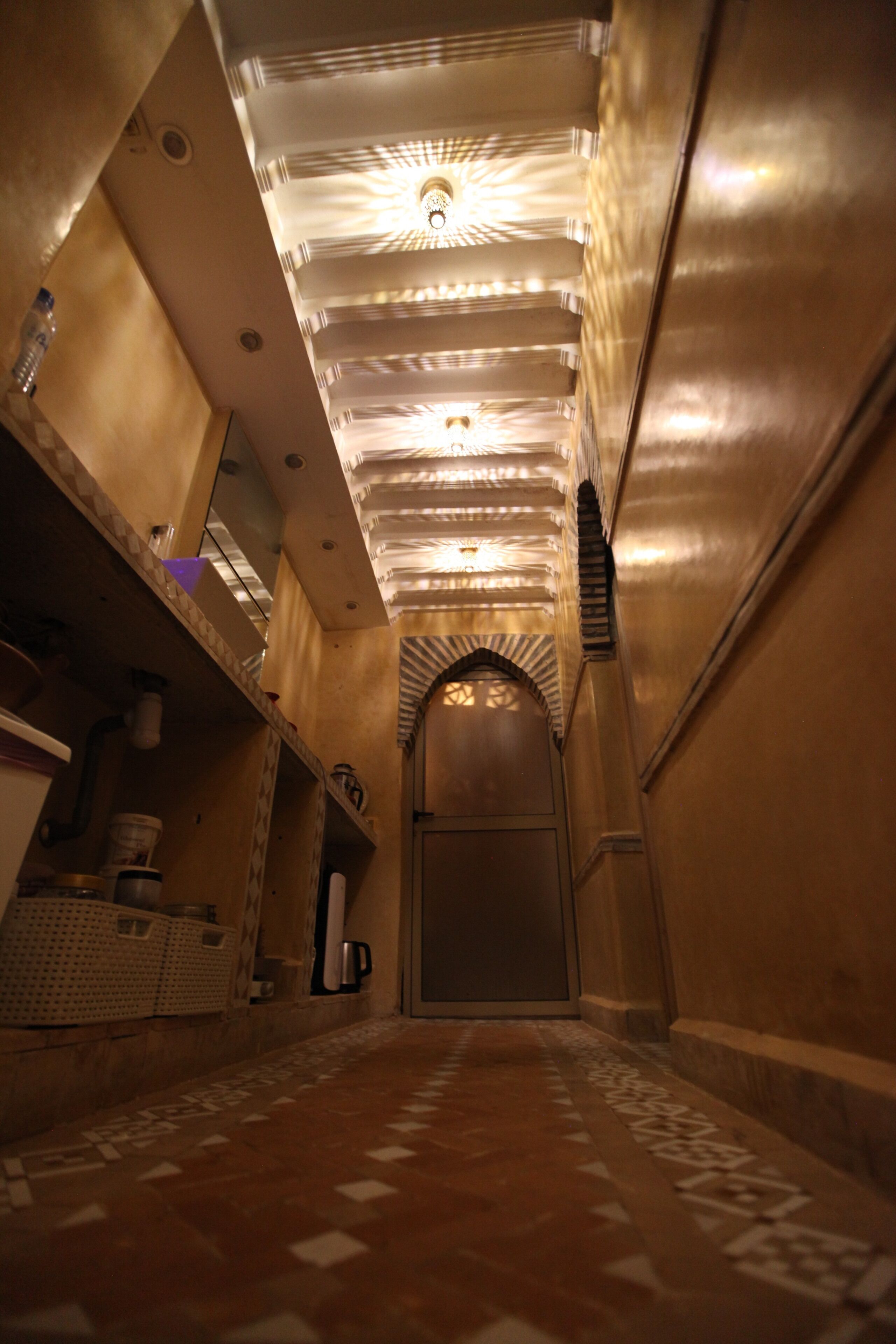 A narrow hallway with patterned tiles, a ceiling with recessed lighting, and a door at the end.