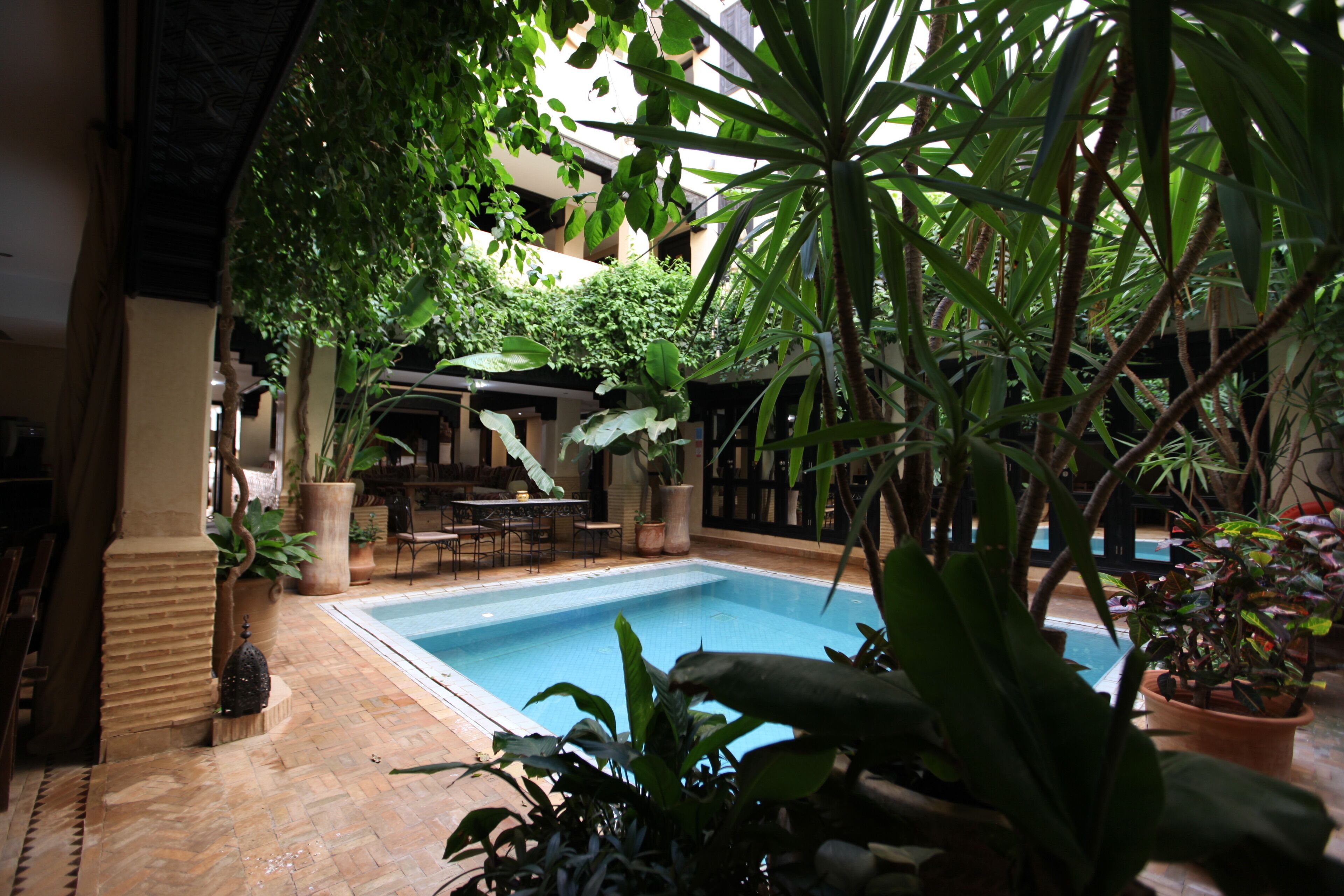 An indoor pool area with a tiled floor, surrounded by potted plants and a seating area with tables and chairs.