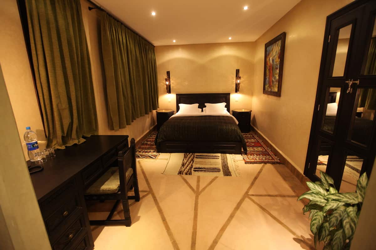 A hotel room with a bed, a desk, a chair, a plant, and a painting on the wall.