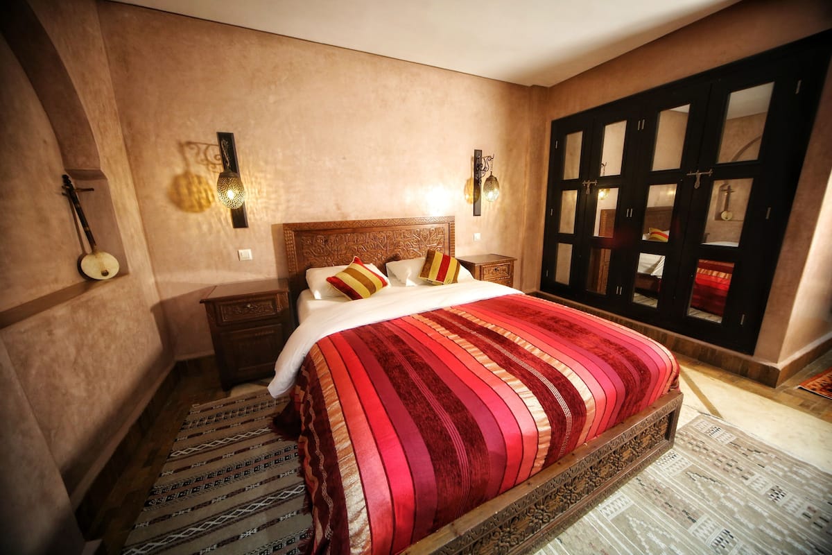 A bedroom with a large bed, a wooden headboard, a wall-mounted lamp, and a patterned rug.