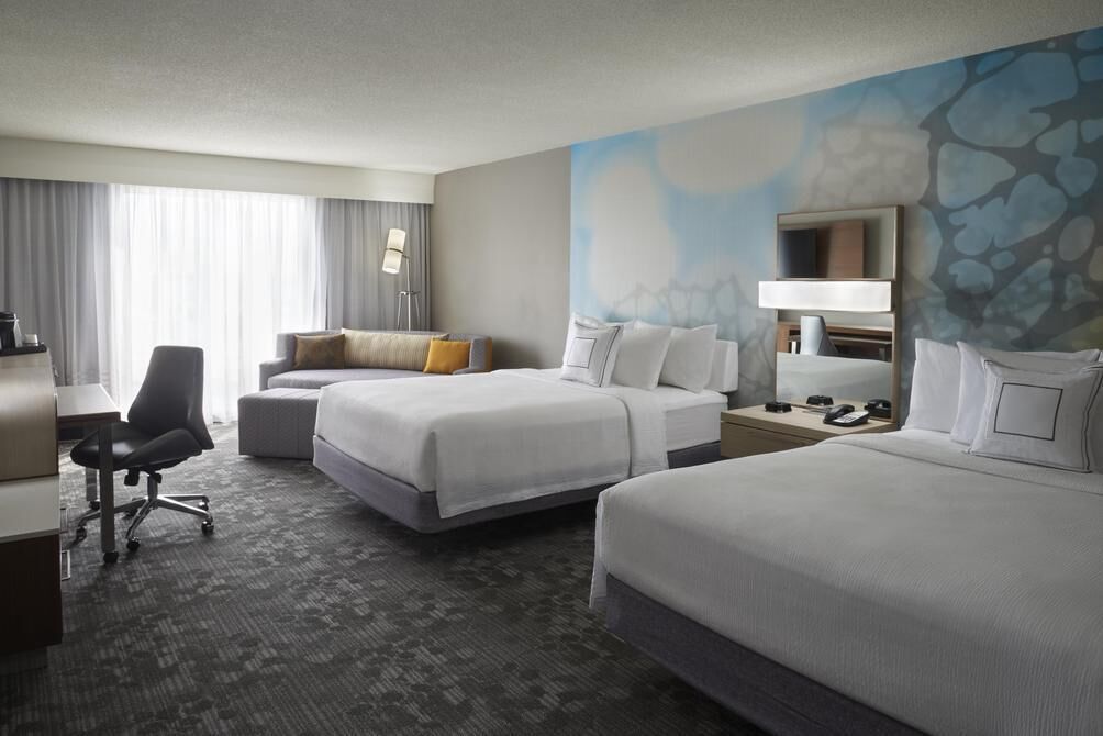 A hotel room with two beds, a desk, a chair, and a large window with curtains.