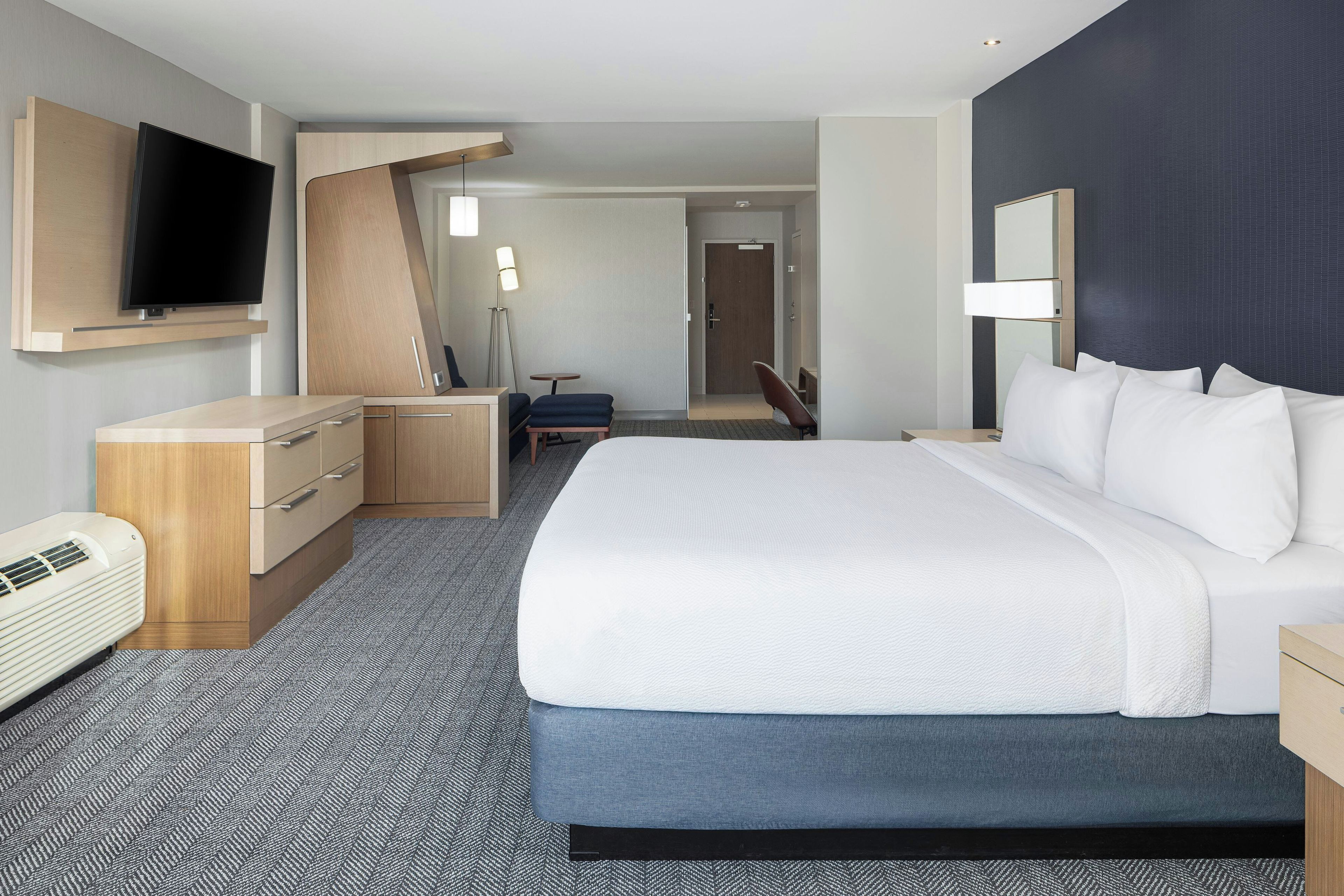 A hotel room with a large bed, a flat-screen TV mounted on the wall, a desk with a chair, a small table, and a lamp.