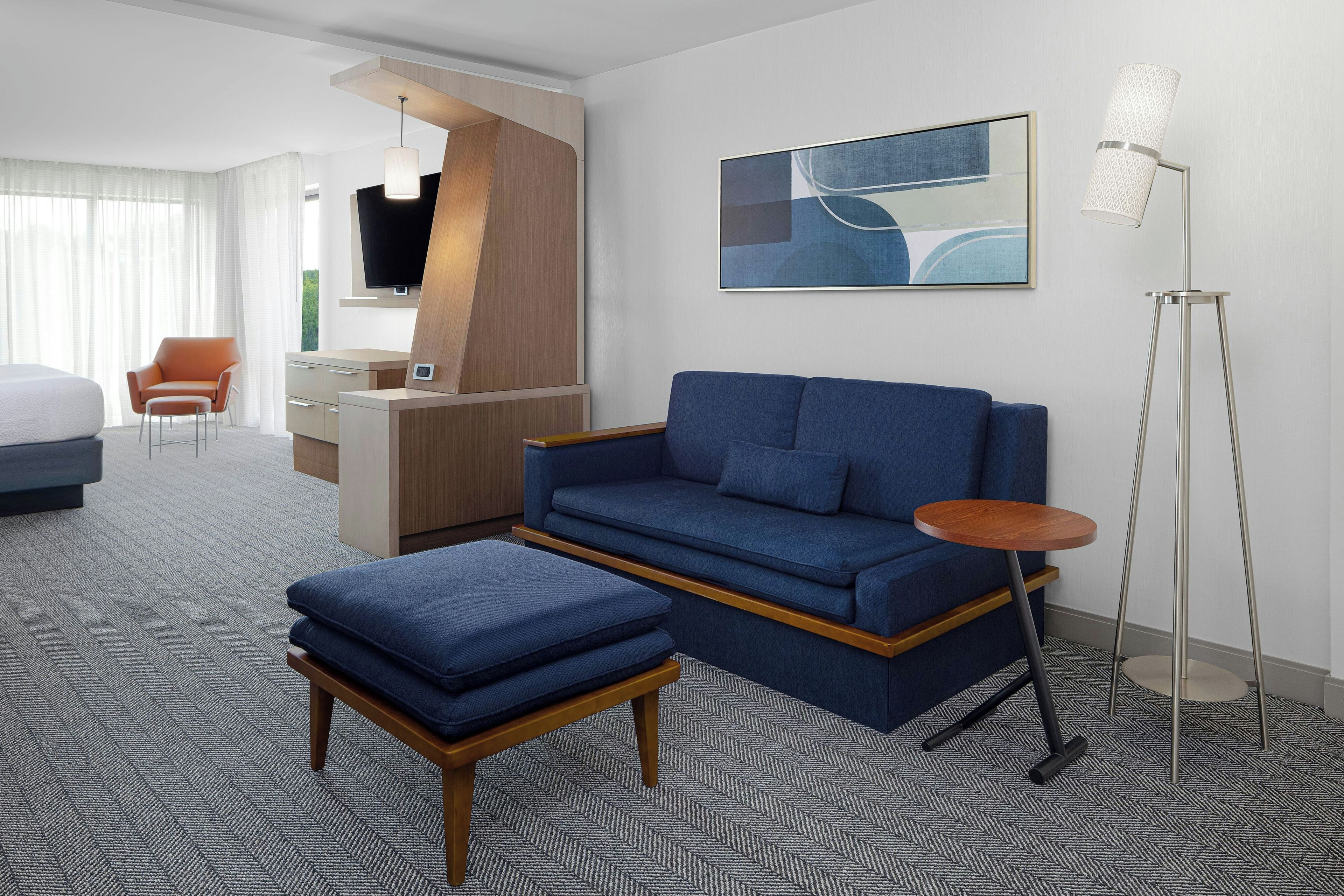 A modern hotel room with a sofa, ottoman, desk, chair, and a floor lamp.