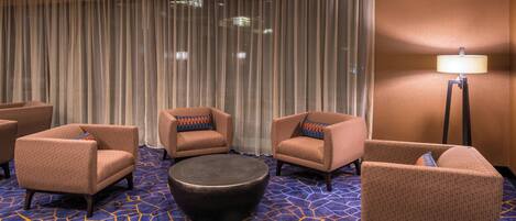 Habitación de hotel con alfombra azul estampada, cuatro sillones, un banquito redondo y una lámpara de pie.