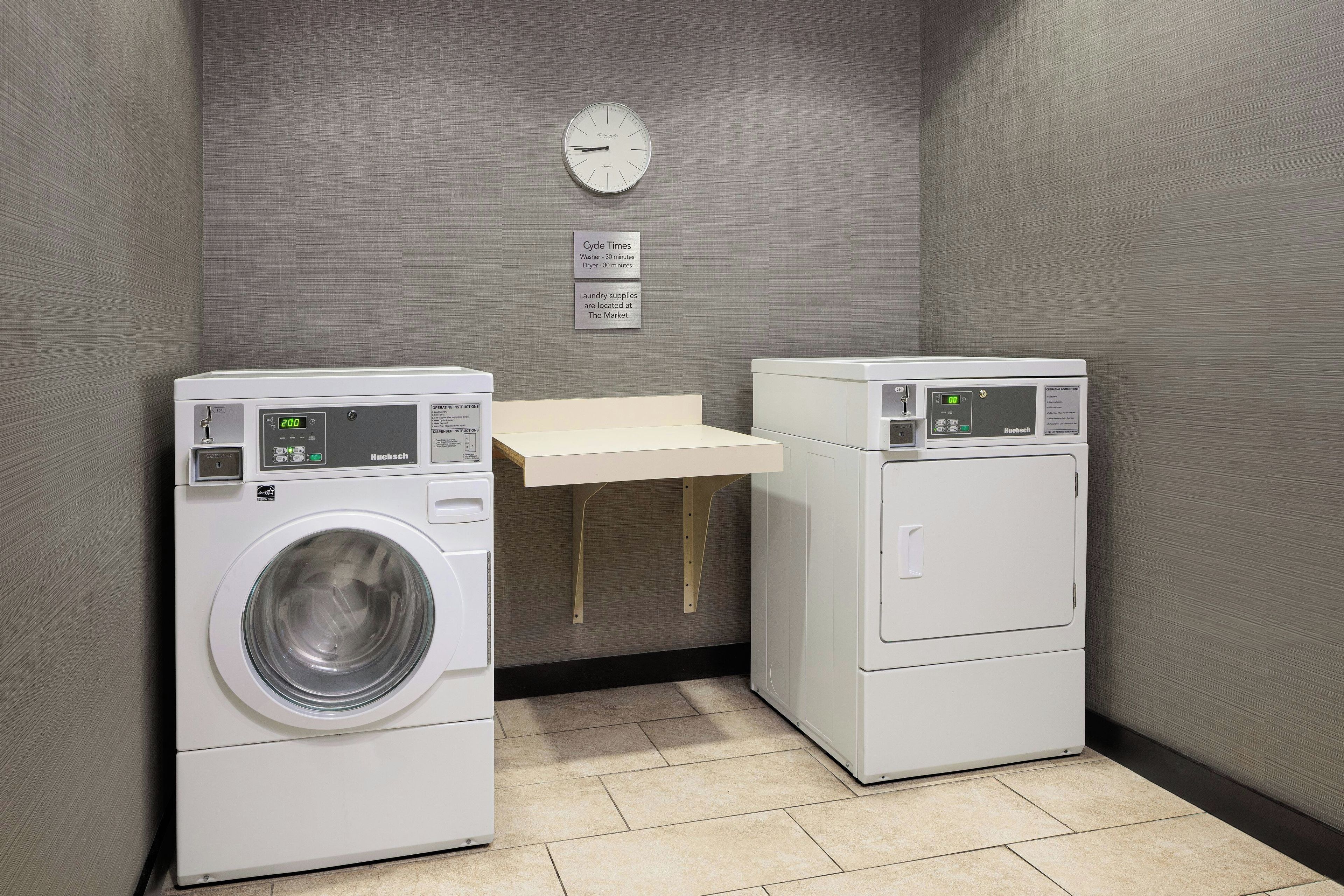 A laundry room with two white front-loading washing machines, a small wooden table, and a wall clock.