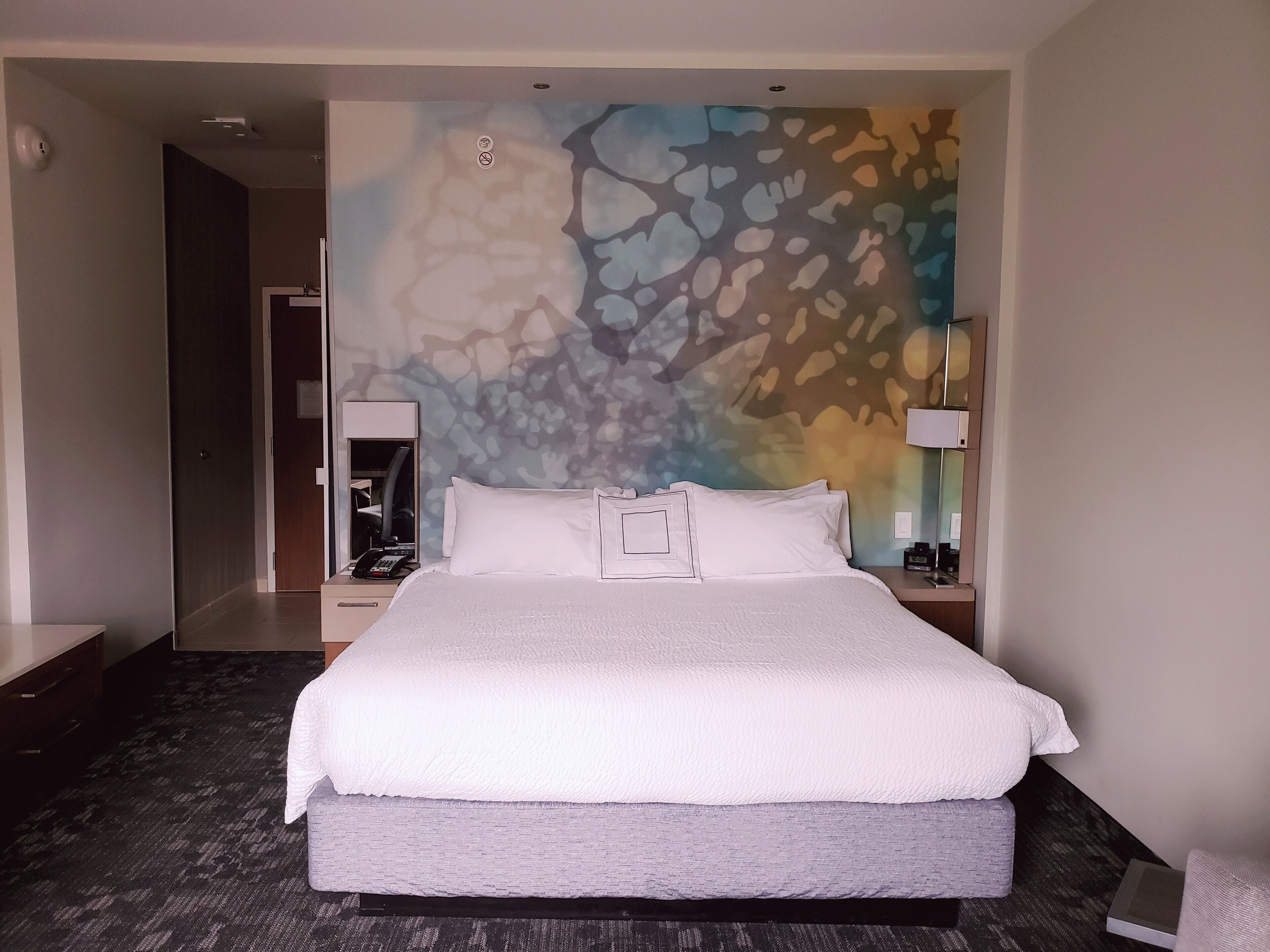 A hotel room with a large bed, a nightstand with a lamp, a wall-mounted telephone, and a door leading to another room.