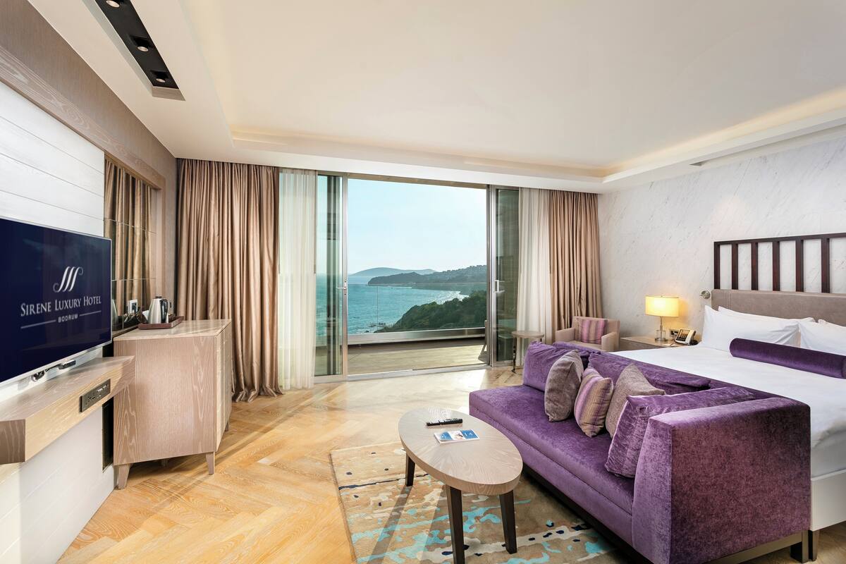 A hotel room with a large TV, a sofa, a bed, a small table, and a view of the sea.