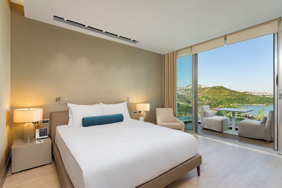 A hotel room with a large bed, two bedside tables with lamps, a chair, and a balcony with a table and chairs.