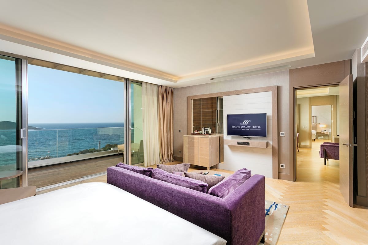 A modern hotel room with a large balcony, a sofa, a TV, and a wooden cabinet.