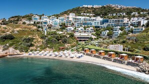 Un resort costero con edificios de varios pisos, una playa con tumbonas y una vista despejada del mar.