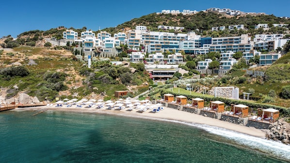 A coastal resort with multiple-story buildings, a beach with sun loungers, and a clear view of the sea.
