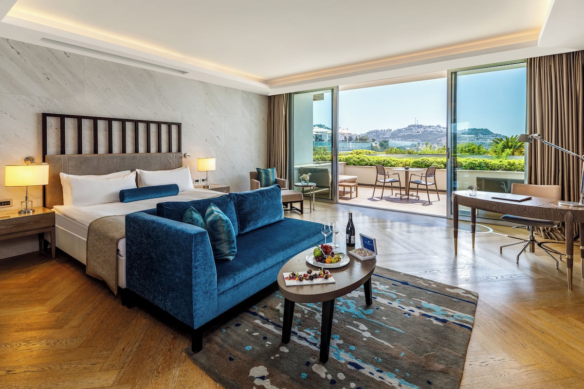 A modern hotel room with a large bed, a blue sofa, a dining area with a table and chairs, and a balcony with a city view.