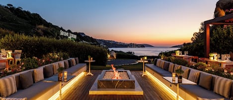 A deck with a fire pit, seating, and tables with candles, overlooking a body of water and hills.