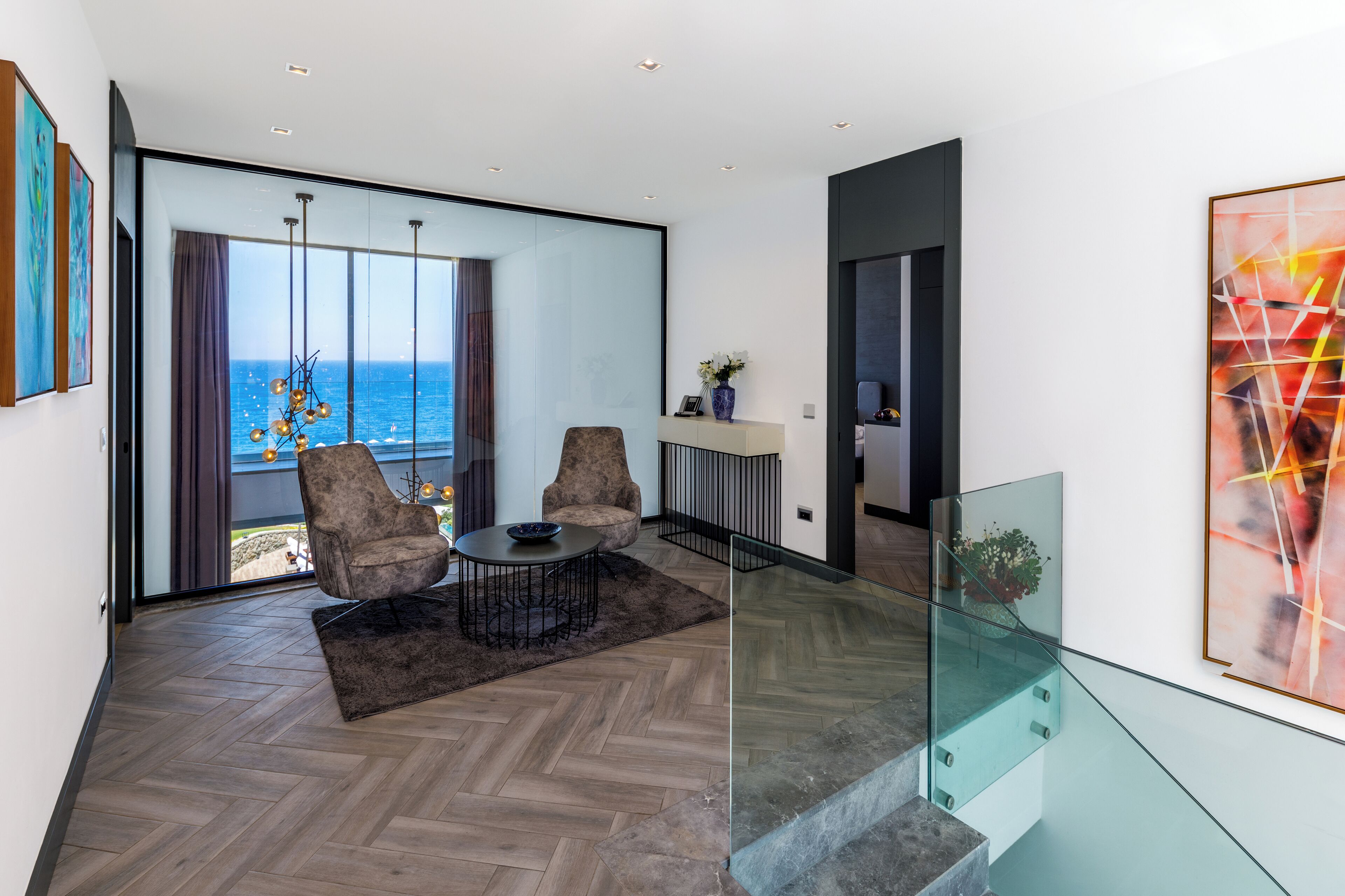 Ein modernes Wohnzimmer mit einem großen Glasfenster, das auf einen Balkon mit Meerblick führt. Es gibt eine Sitzecke mit zwei Stühlen und einem Tisch, sowie eine Glastrennwand zum Balkon.