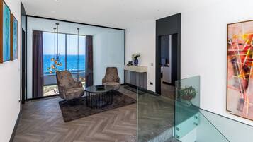 Ein modernes Wohnzimmer mit einem großen Glasfenster, das auf einen Balkon mit Meerblick führt. Es gibt eine Sitzecke mit zwei Stühlen und einem Tisch, sowie eine Glastrennwand zum Balkon.