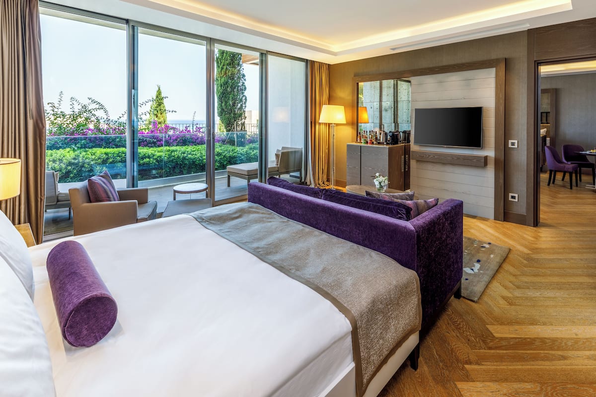 A modern hotel room with a large bed, a sofa, a TV, and a balcony with a view of greenery.