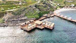 Vista aérea de una propiedad frente al mar con estructuras flotantes de madera, una playa y una colina con vegetación.