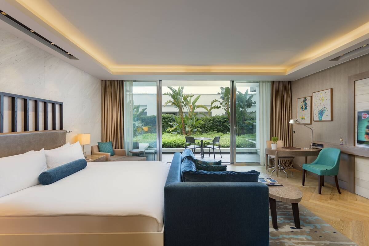 A hotel room with a large bed, a desk, a chair, and a sofa. There is a view of the outdoors through the windows.