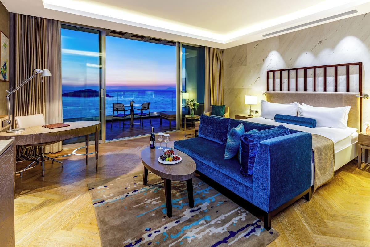 A hotel room with a large bed, a sofa, a dining table, and a view of the ocean.