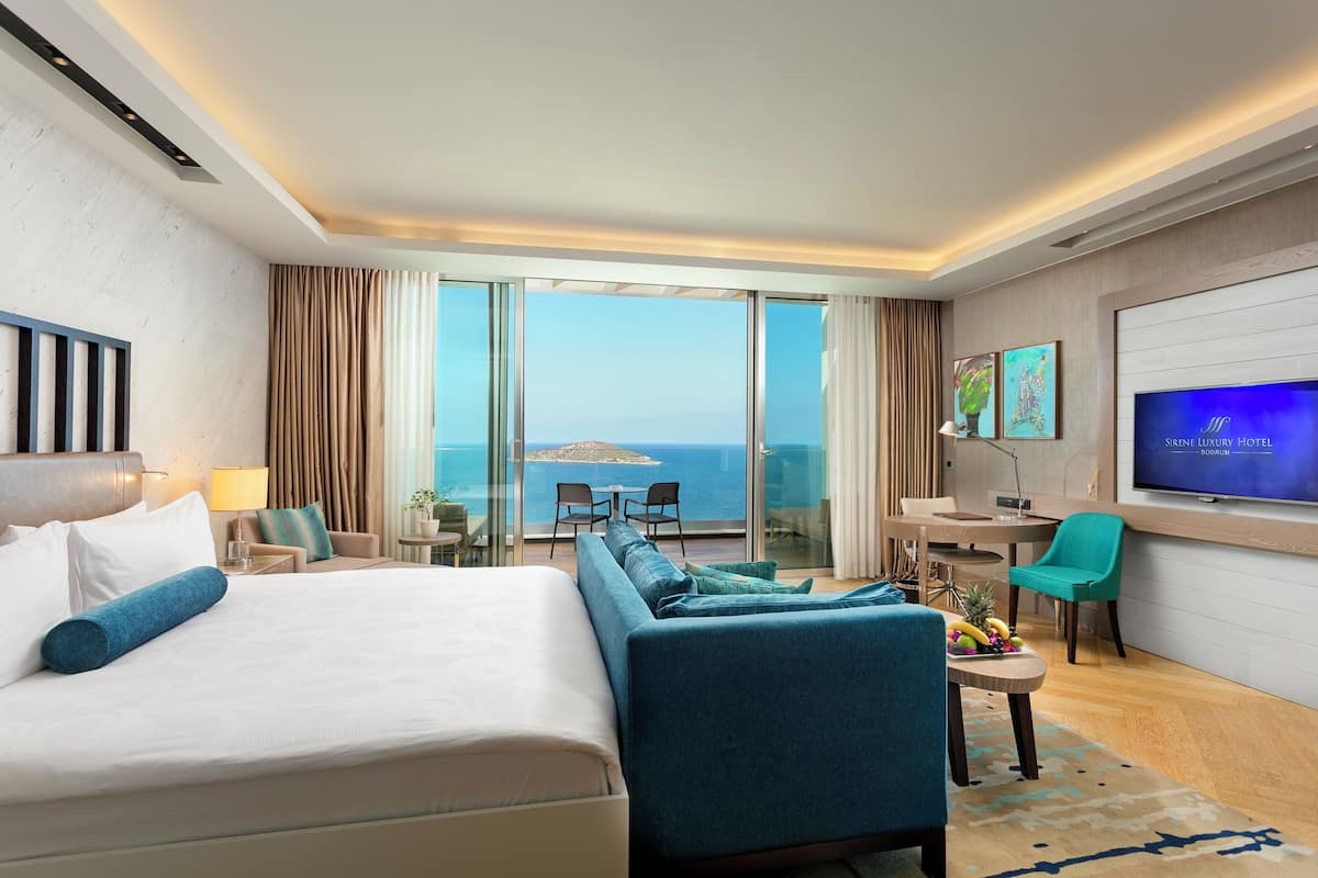 A modern hotel room with a large bed, a sofa, a TV, and a balcony with a view of the sea.