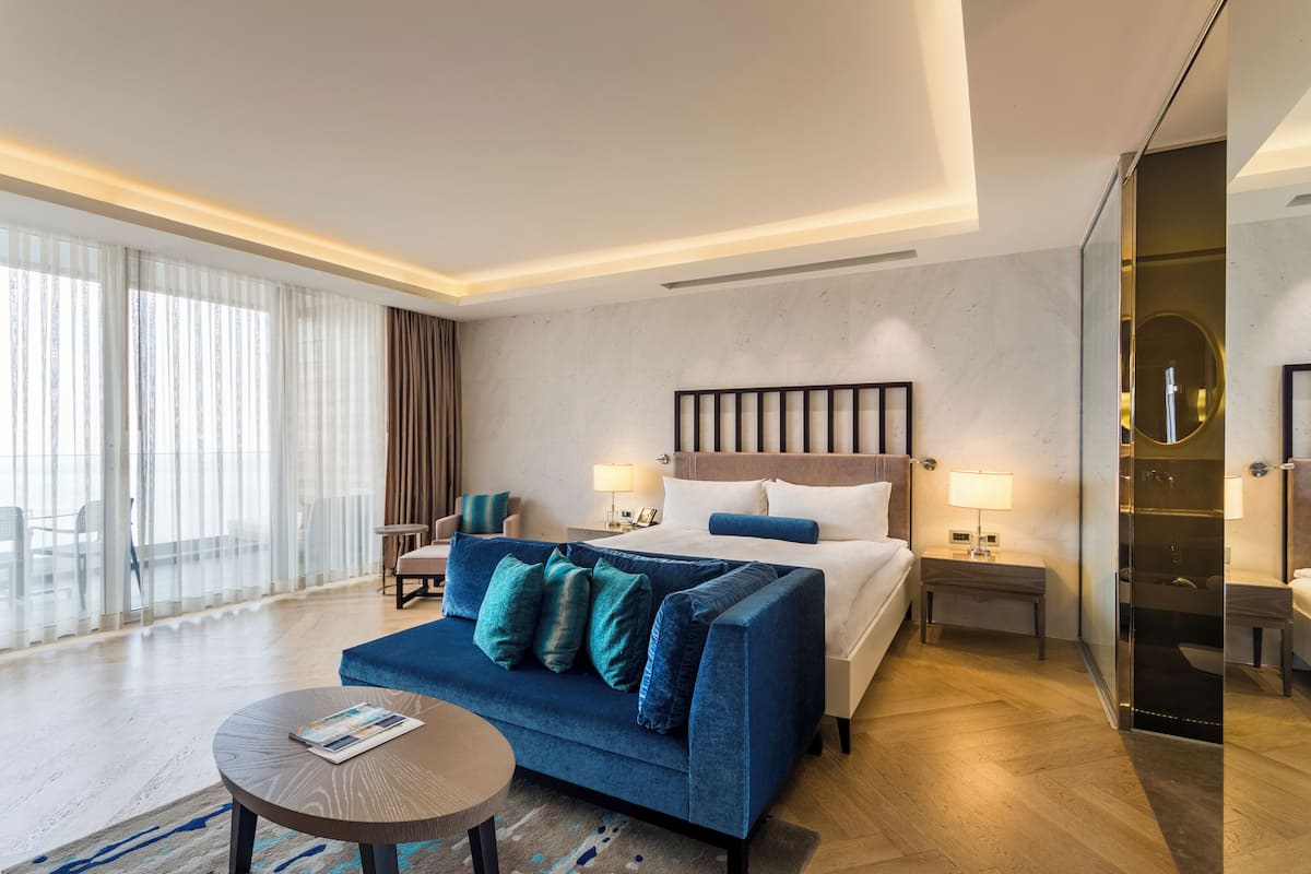 A hotel room with a bed, a blue sofa, a round table, a glass shower enclosure, and a large window with curtains.