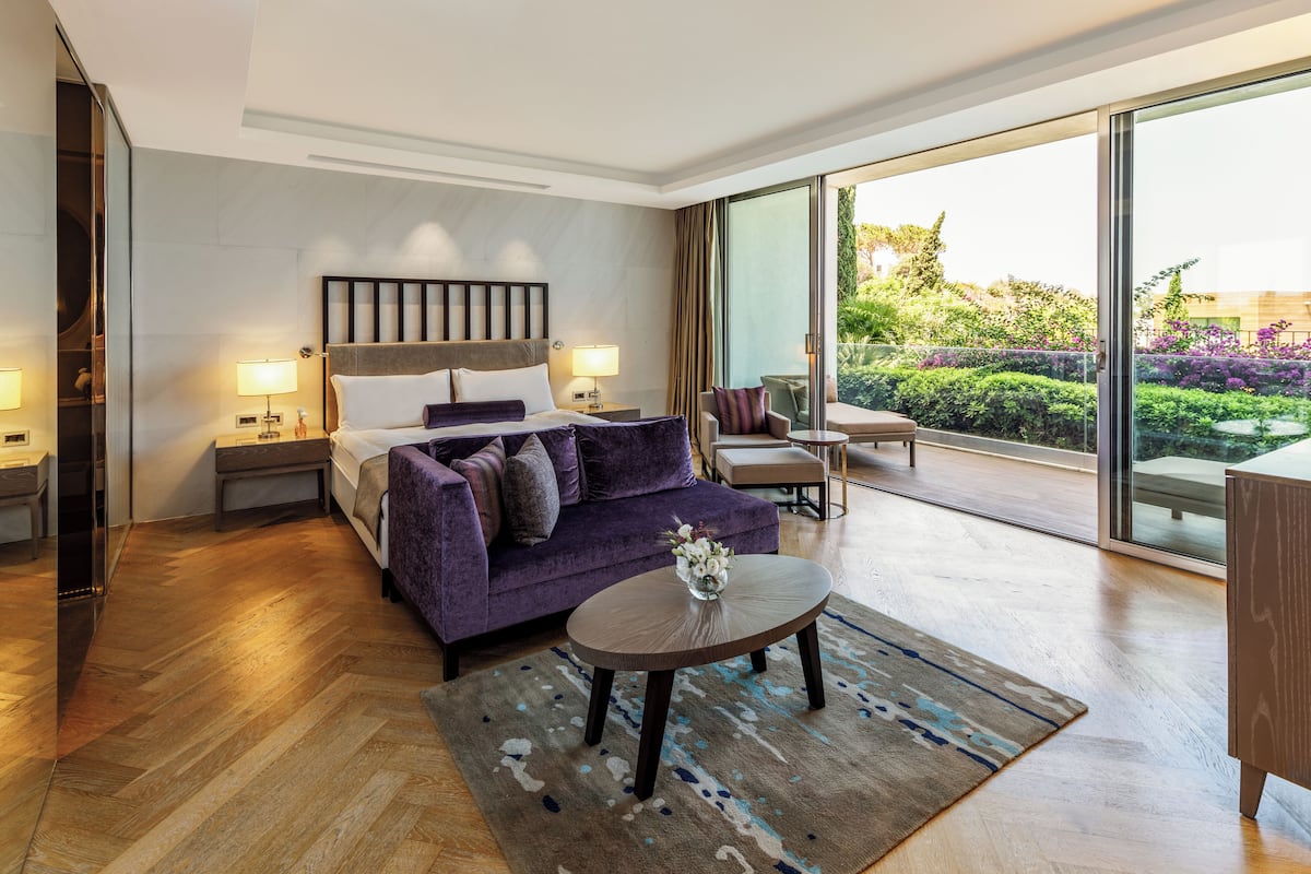 A modern hotel room with a large bed, a sofa, a round coffee table, and a balcony with a view of greenery.