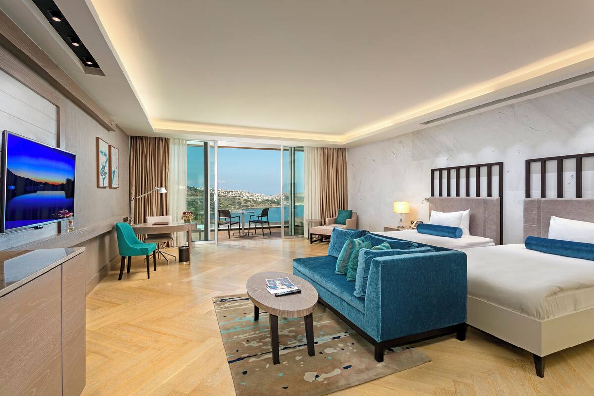 A modern hotel room with a sofa, two beds, a TV, a desk, and a view of the city.