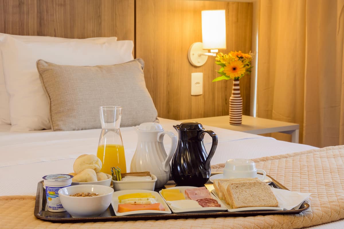 A neatly arranged breakfast tray on a bed with a wooden headboard, including a bowl of cereal, a jug of orange juice, a white pitcher, a black coffee pot, and a plate with eggs, ham, and bread.