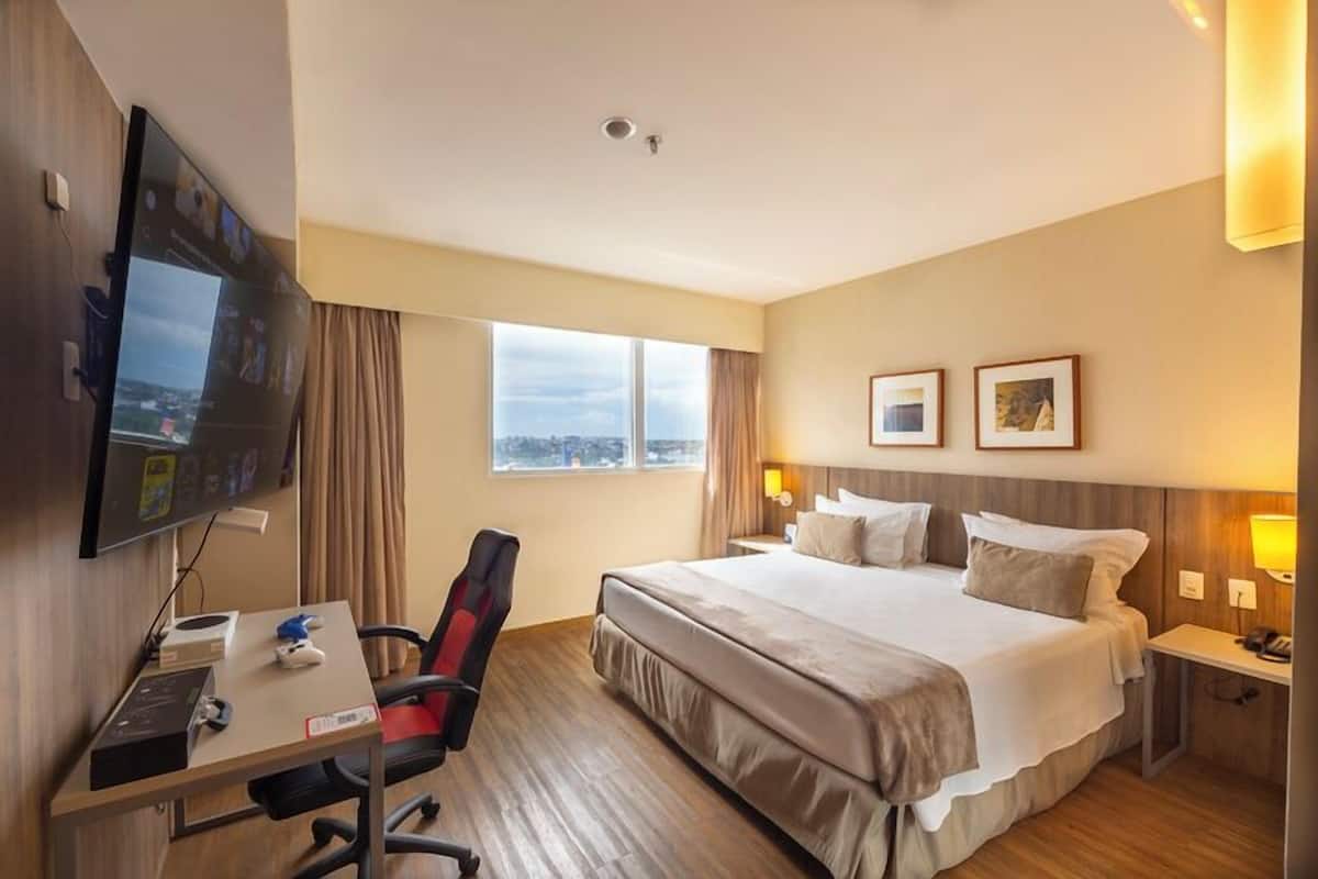 A hotel room with a large bed, a desk with a chair, a flat-screen TV mounted on the wall, and a window with curtains.
