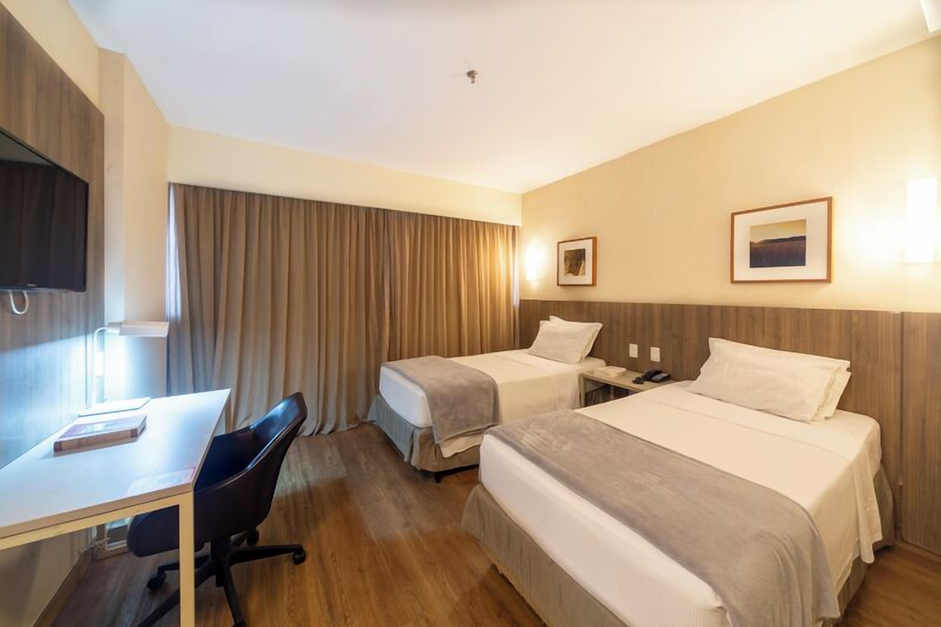 A hotel room with two beds, a desk, a chair, a TV, and a window with curtains.