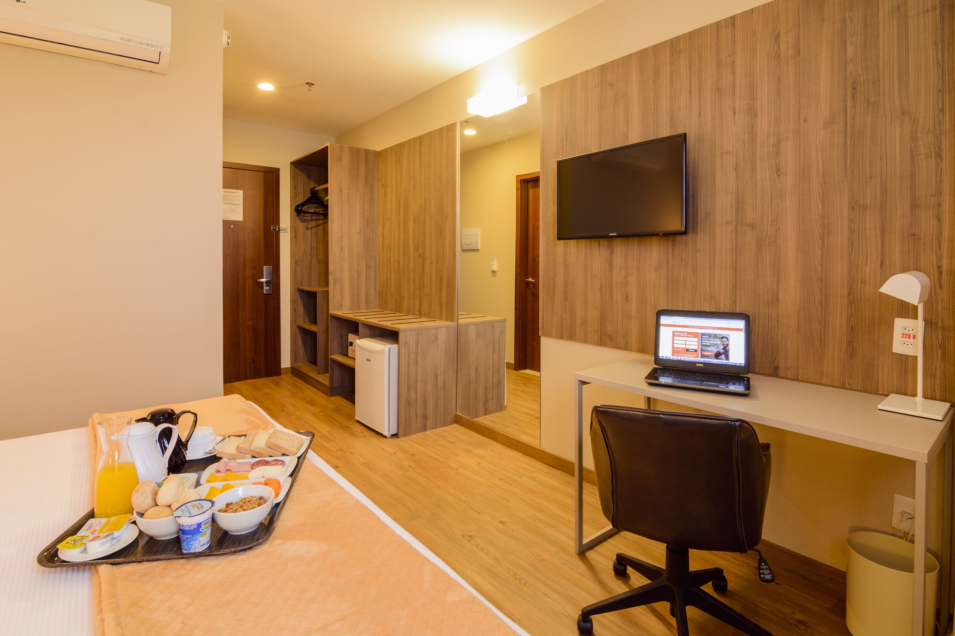 Une chambre d’hôtel avec un plancher en bois, un bureau avec un ordinateur portable, un téléviseur fixé au mur, une petite kitchenette et un plateau de petit-déjeuner sur le lit.