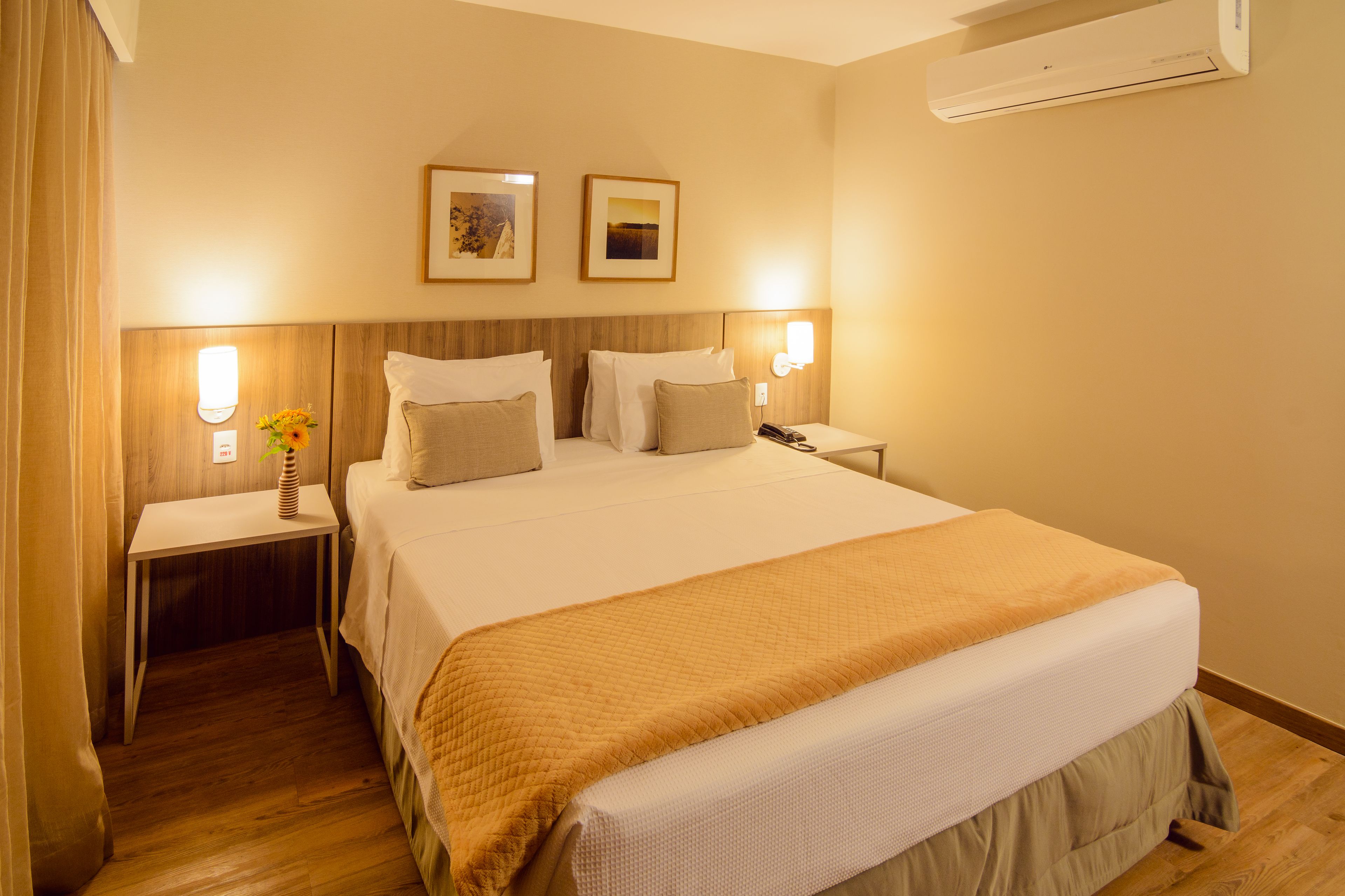 A hotel room with a large bed, bedside tables, a nightstand, and an air conditioning unit.