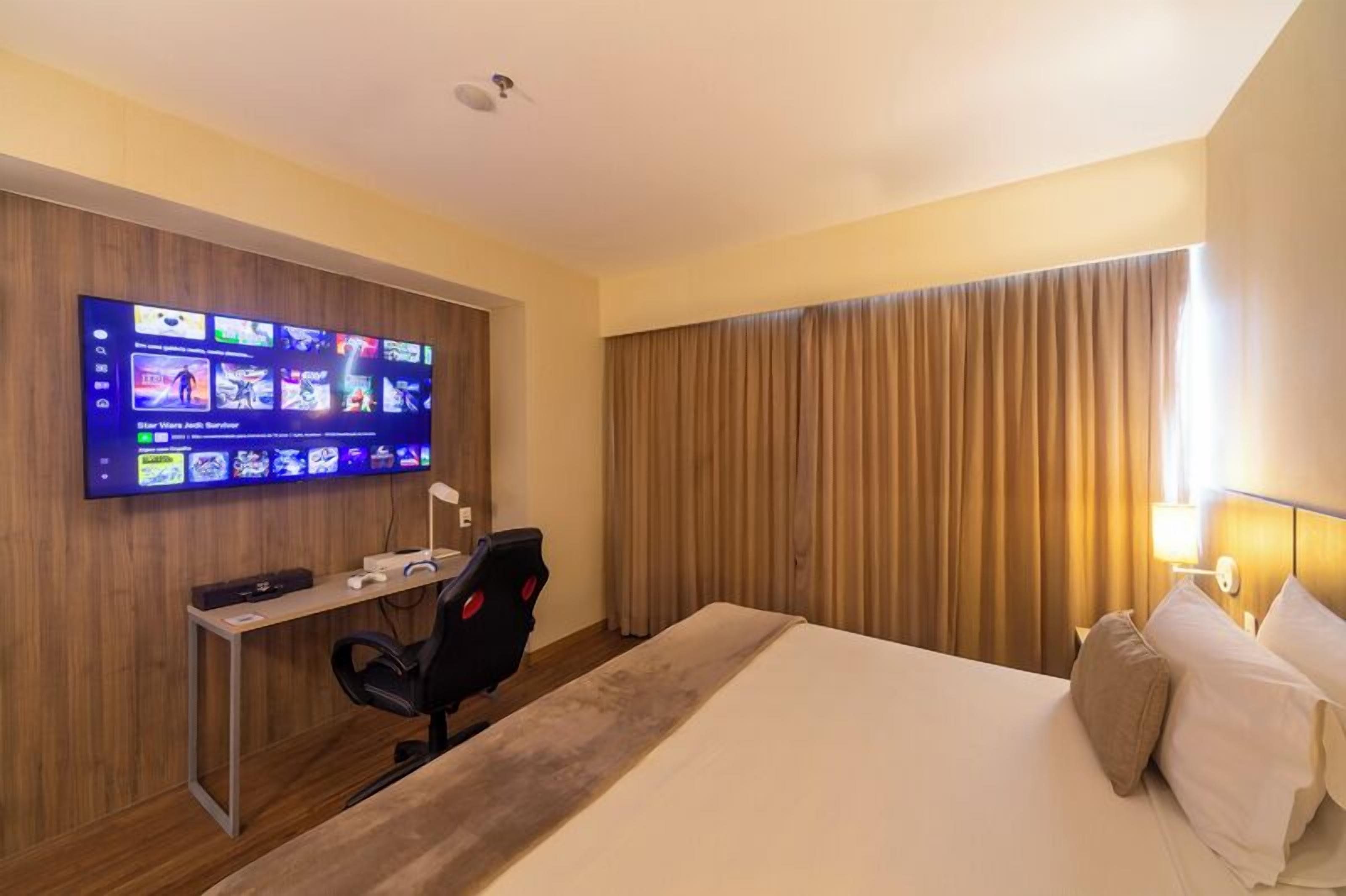 A hotel room with a large flat-screen TV mounted on the wall displaying various games, a desk with a computer and headphones, a comfortable bed with pillows, and a large window with curtains.