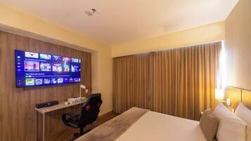 A hotel room with a large flat-screen TV mounted on the wall displaying various games, a desk with a computer and headphones, a comfortable bed with pillows, and a large window with curtains.