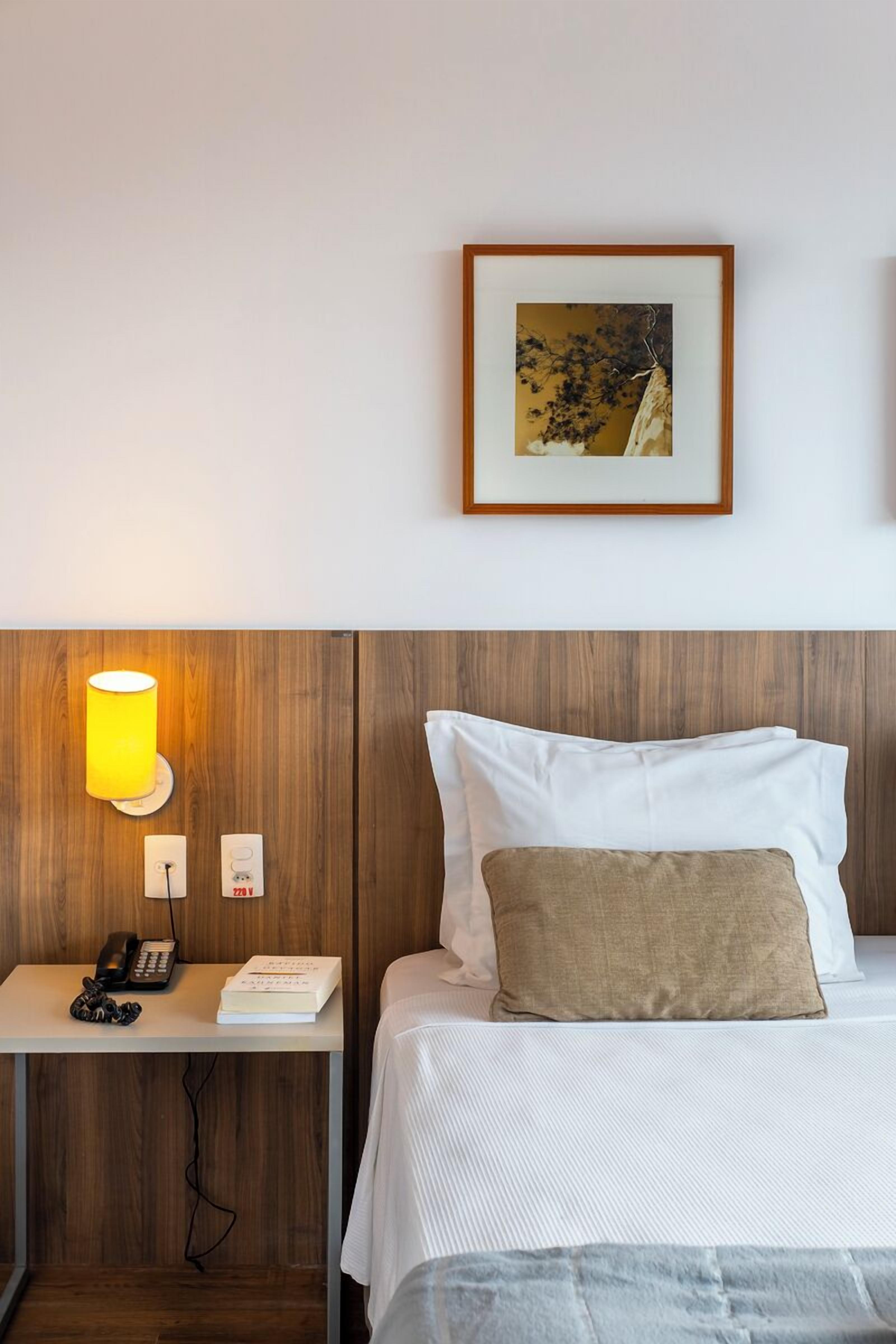 Habitación de hotel con cabecera de madera, mesita de noche con lámpara, teléfono, libro y un cuadro enmarcado en la pared.