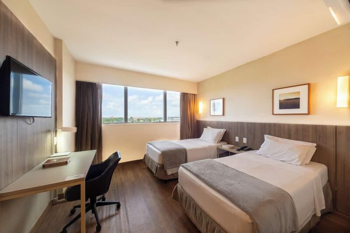 A hotel room with two beds, a desk, a chair, a TV, and a window with a view.