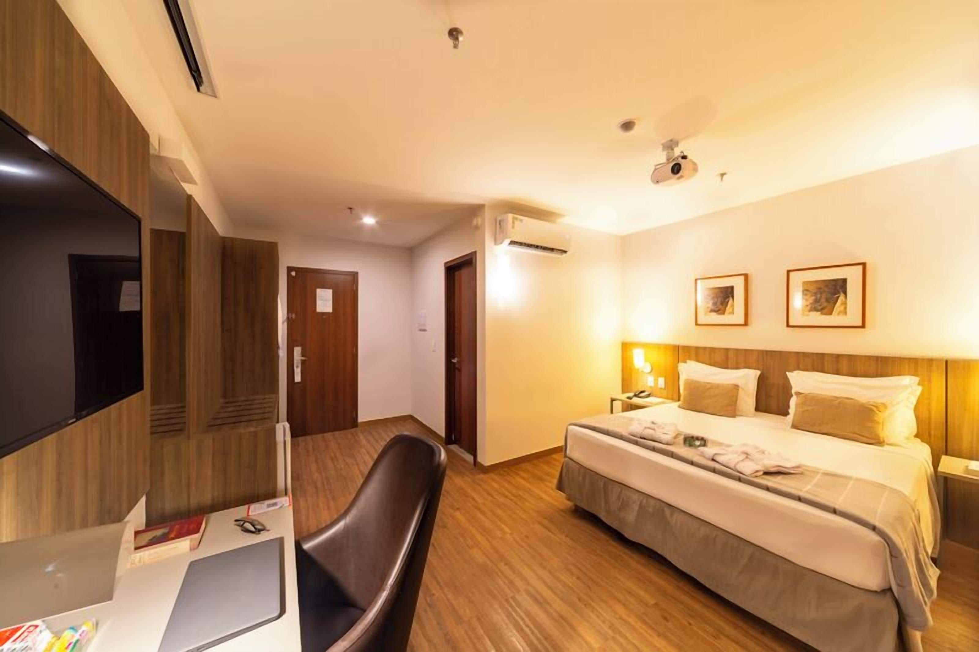 A hotel room with a bed, a desk, a chair, a television, and a ceiling fan.