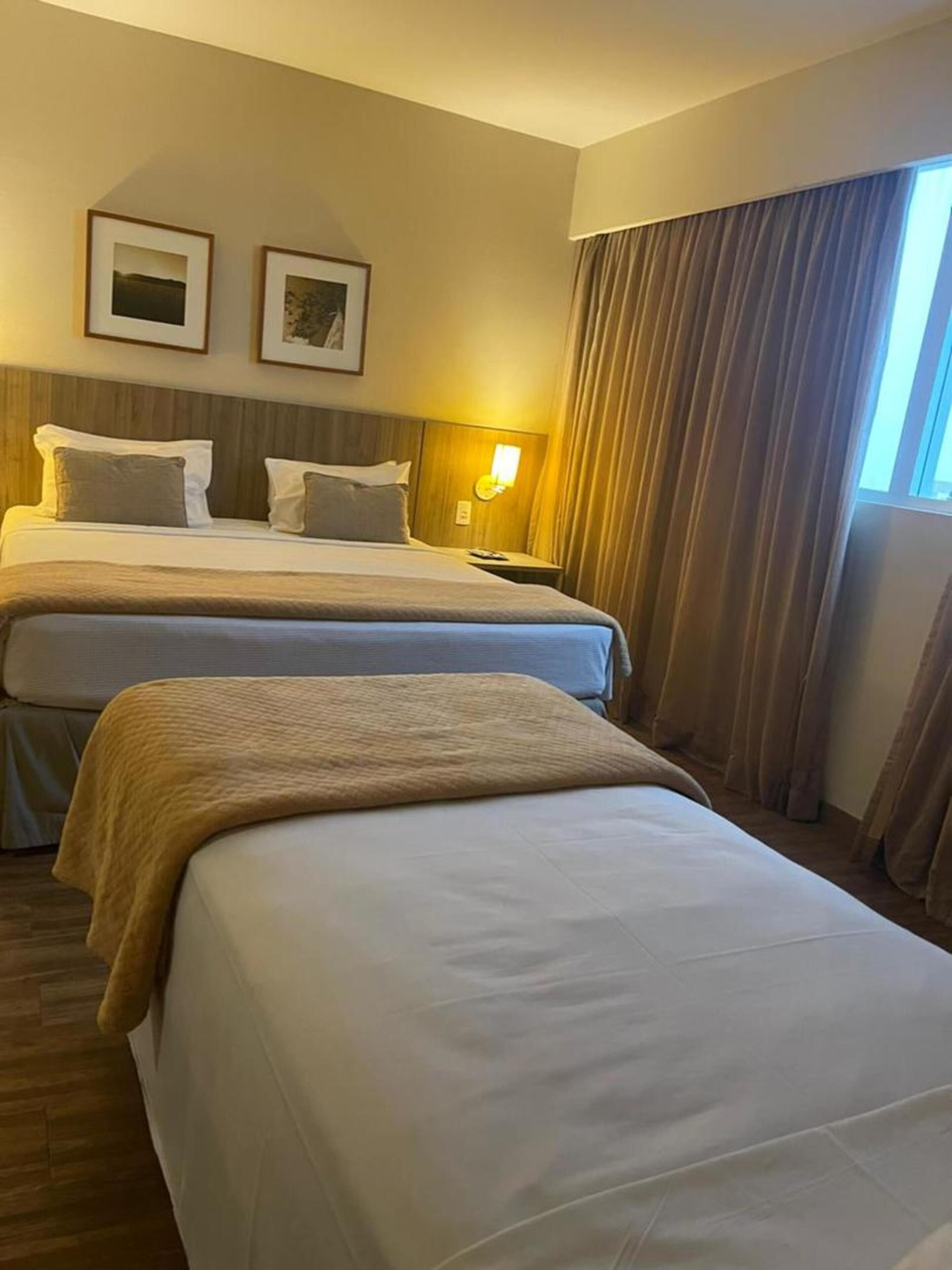 A hotel room with two beds, a nightstand, a lamp, and a large window with curtains.