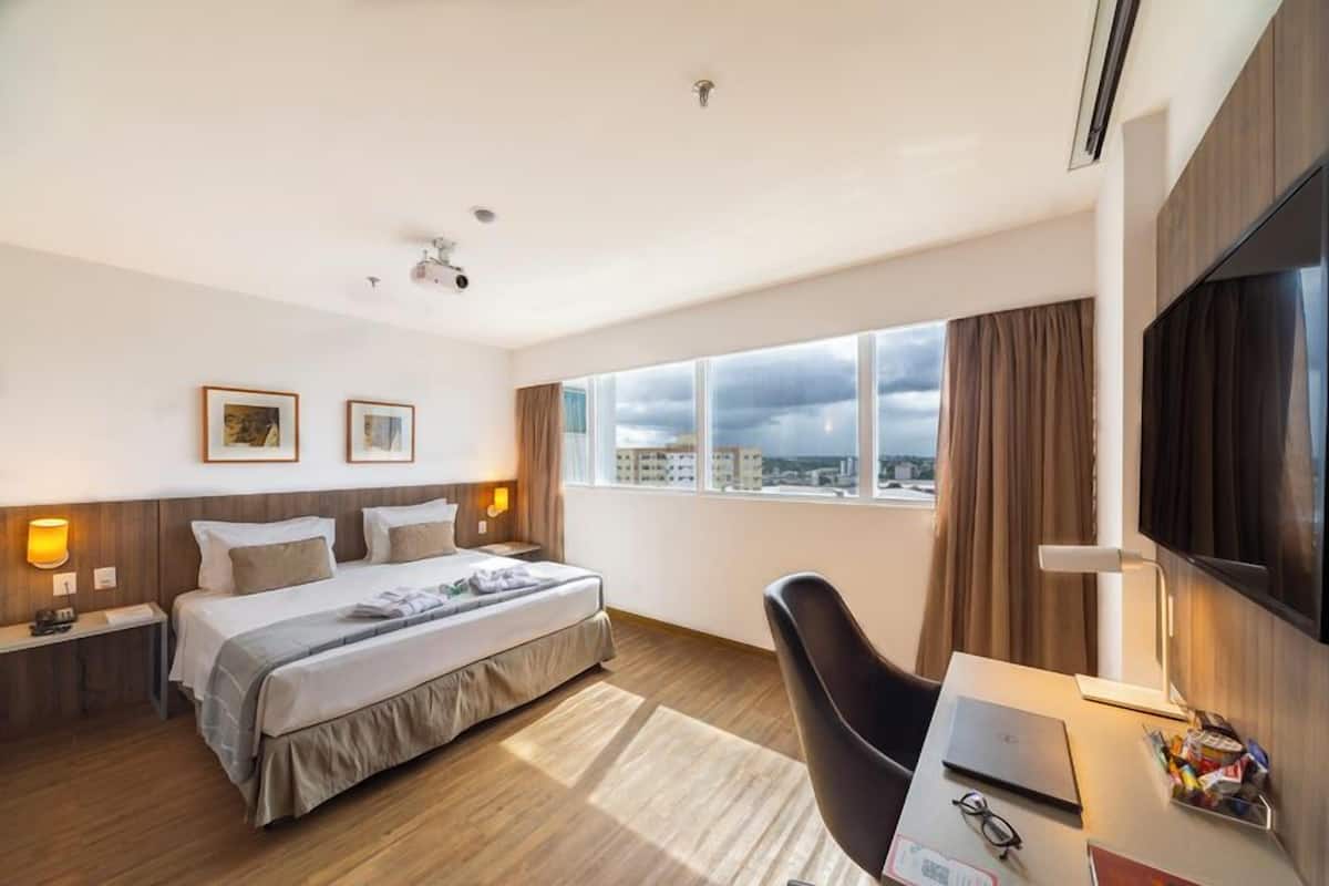A modern hotel room with a large bed, a desk with a laptop, and a view of the city through large windows.
