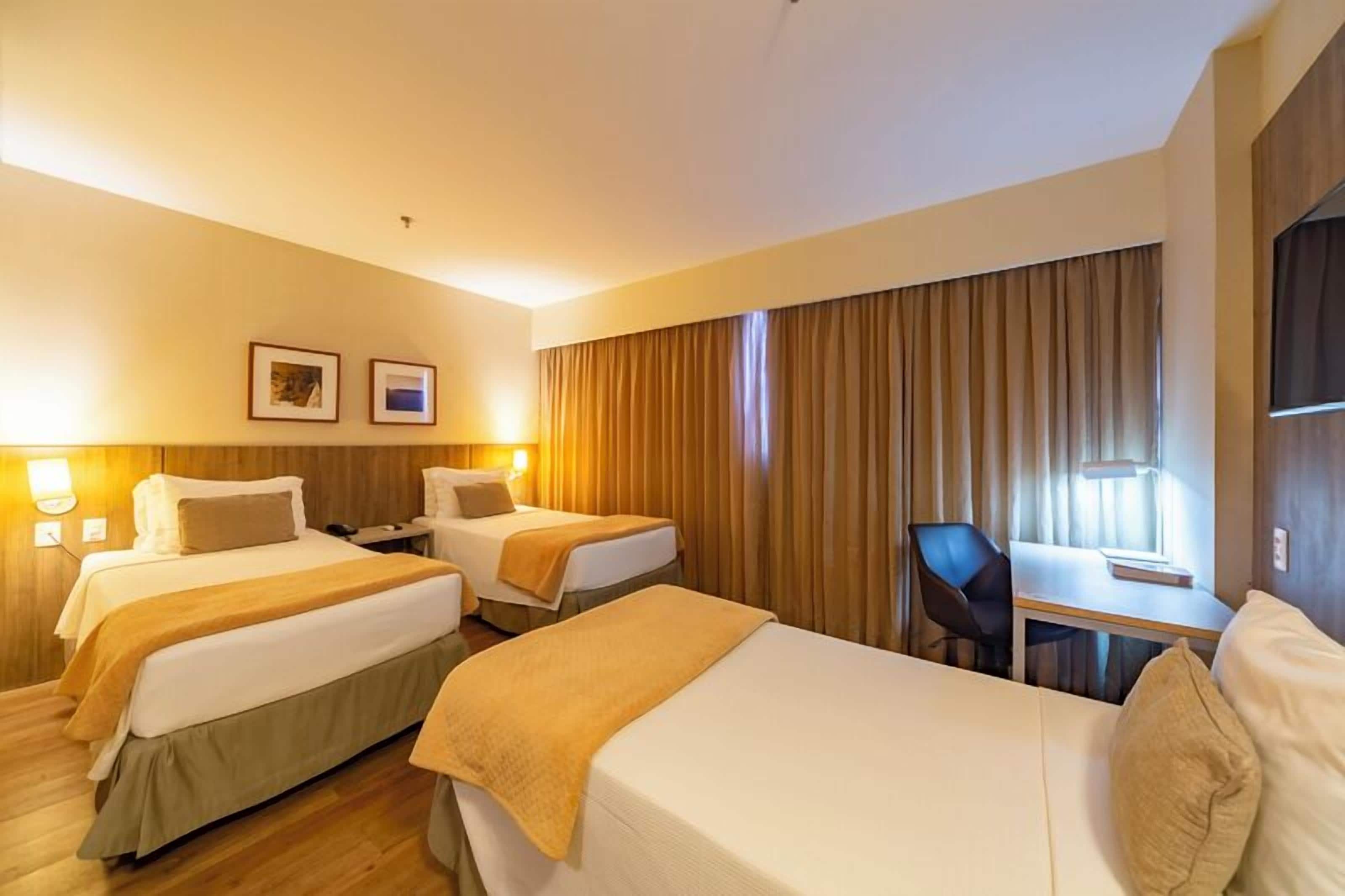 A hotel room with two beds, a desk, and a chair. There are two wall-mounted lamps and a window with curtains.