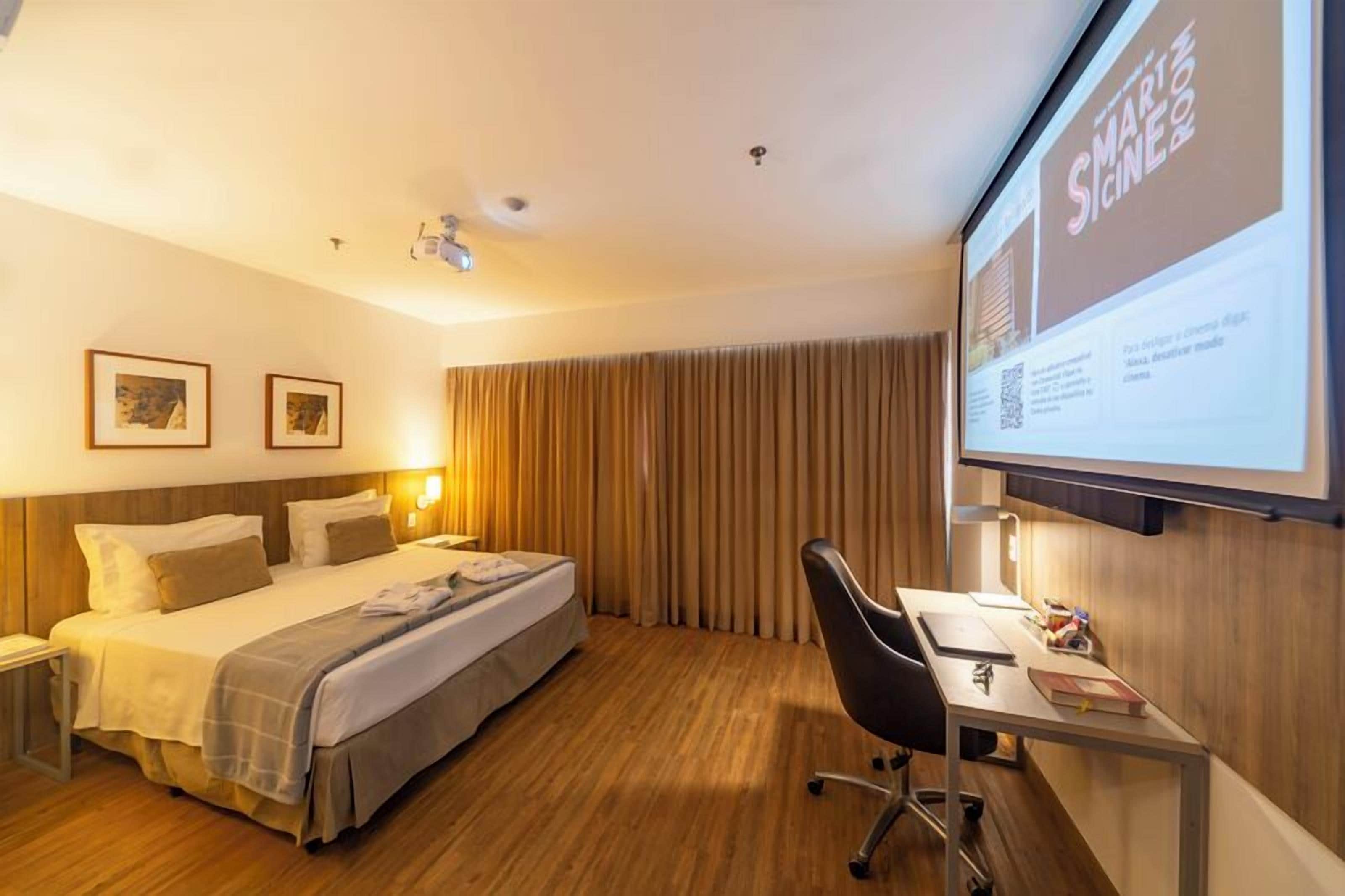 A hotel room with a bed, a desk, a chair, a projector screen, and curtains.