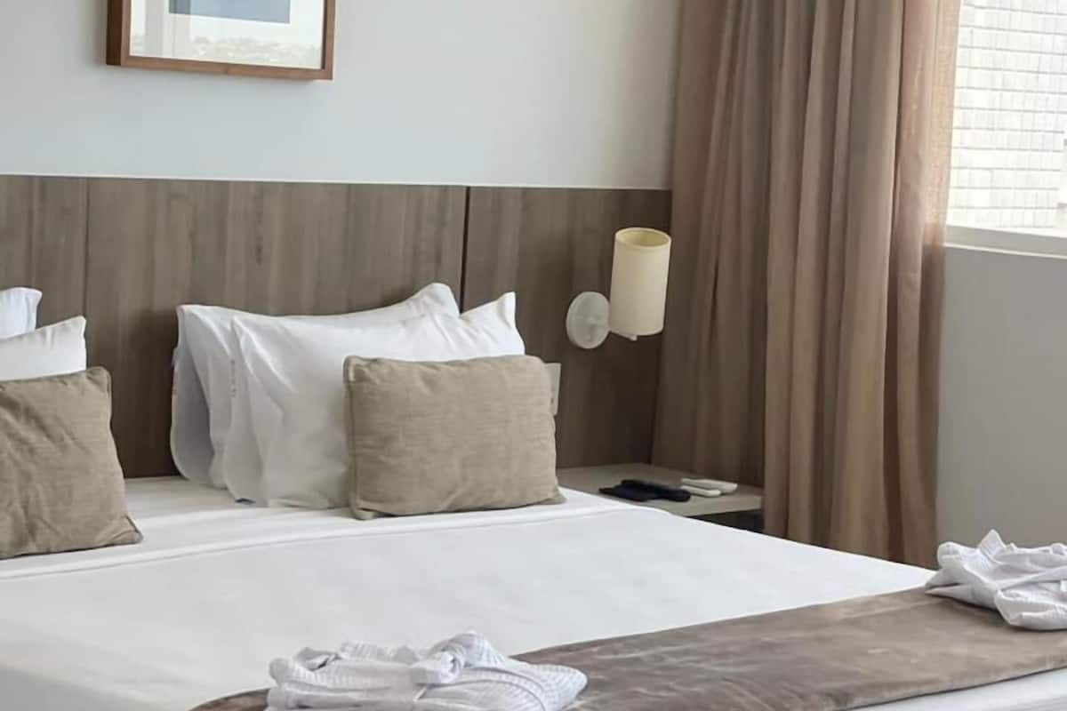 A neatly made bed with a brown throw blanket and folded towels, a wooden headboard, a framed picture on the wall, and a bedside lamp.