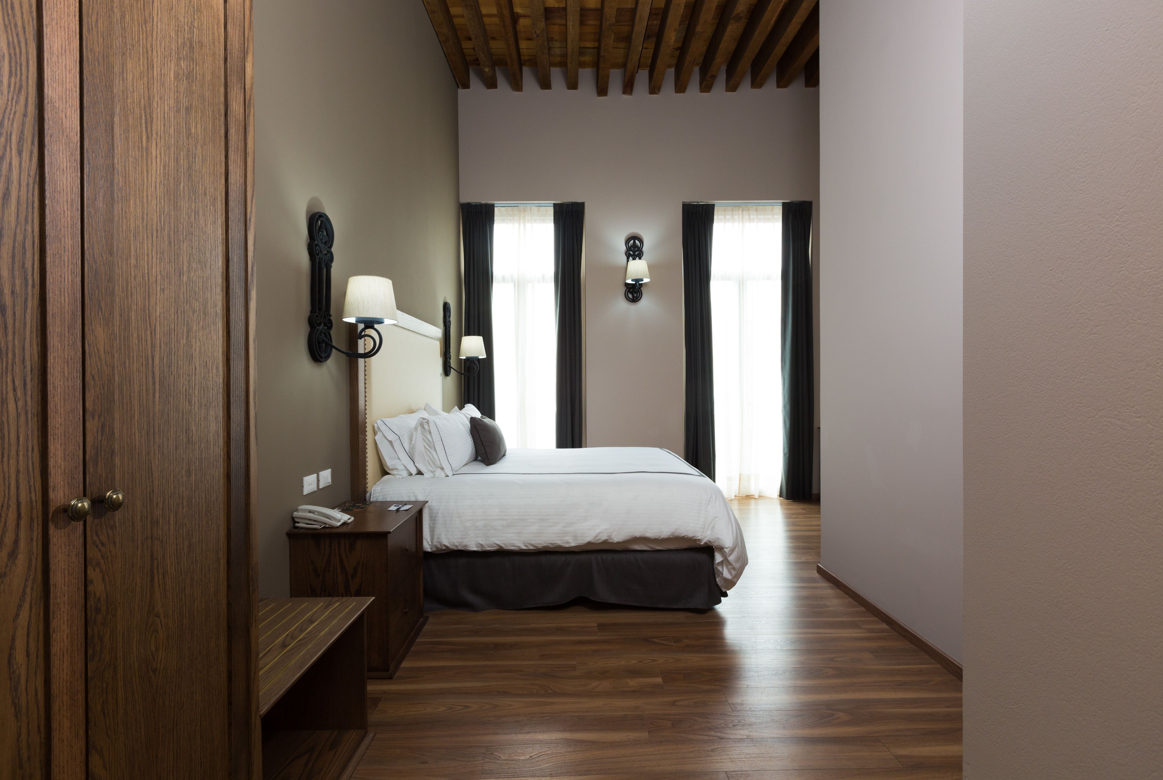 A bedroom with a wooden floor, a bed with a white duvet, a nightstand, a lamp, and a wall-mounted light fixture.