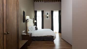 A bedroom with a wooden floor, a bed with a white duvet, a nightstand, a lamp, and a wall-mounted light fixture.