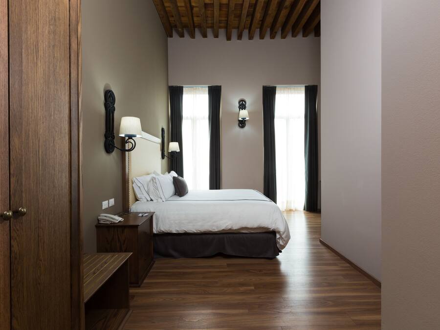A bedroom with a wooden floor, a bed with a white duvet, a nightstand, a lamp, and a wall-mounted light fixture.